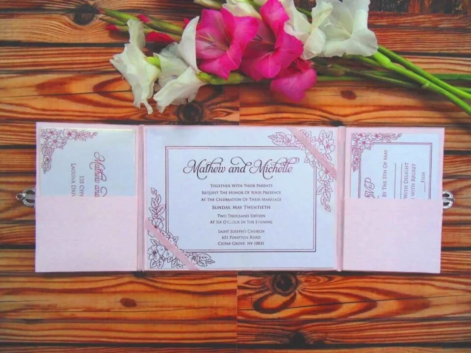 An open pale blush pink tri-fold wedding invitation suite laid flat on a rustic wood-grain surface, topped with a bouquet of vibrant pink and white gladiolus flowers. Center Panel: The main invitation card is white with a thin pink border and delicate pink floral illustrations in the corners. It features the name.the card is held in place by diagonal pink ribbons. Left and Right Panels: Each side features a blush pink pocket. The left pocket holds a details card, and the right pocket holds an RSVP card, both matching the floral theme and pink-on-white color scheme.