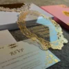 An angled close-up view of a luxury clear acrylic wedding invitation and coordinating stationery, set against a textured surface. The highlight of the suite is: An oval-shaped clear acrylic invitation featuring an intricate, laser-etched gold scrollwork border. The formal event details are printed in gold at the center of the plaque. The invitation rests partially atop a matching white invitation box with a gold-bordered lid. In the foreground, a gold-foiled RSVP card** is visible, featuring the initials and elegant floral accents in shimmering gold. In the background, a pink invitation folio with a gold central monogram adds a soft pop of color to the arrangement. The scene is captured in natural sunlight, creating soft shadows and emphasizing the high-shine, metallic finish of the gold elements.