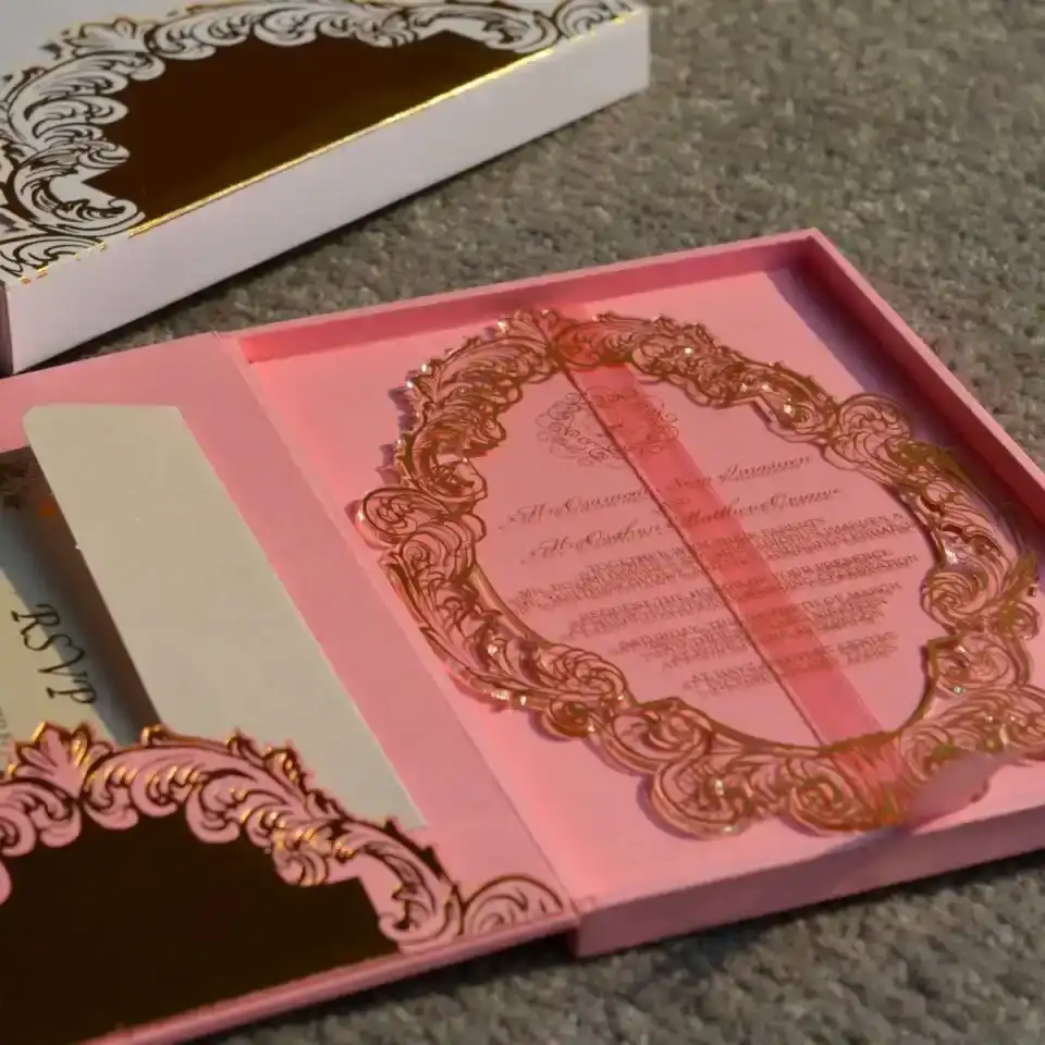 An angled close-up view of a luxury clear acrylic wedding invitation and coordinating stationery, set against a textured surface. The highlight of the suite is: An oval-shaped clear acrylic invitation featuring an intricate, laser-etched gold scrollwork border. The formal event details are printed in gold at the center of the plaque. The invitation rests partially atop a matching white invitation box with a gold-bordered lid. In the foreground, a gold-foiled RSVP card** is visible, featuring the initials and elegant floral accents in shimmering gold. In the background, a pink invitation folio with a gold central monogram adds a soft pop of color to the arrangement. The scene is captured in natural sunlight, creating soft shadows and emphasizing the high-shine, metallic finish of the gold elements.