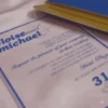A transparent acrylic wedding invitation is shown partially tucked into a textured royal blue envelope. The invitation rests on a draped, light pink satin fabric that creates soft, shimmering reflections on the clear surface.