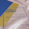 A transparent acrylic wedding invitation is shown partially tucked into a textured royal blue envelope. The invitation rests on a draped, light pink satin fabric that creates soft, shimmering reflections on the clear surface.