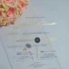A transparent acrylic wedding invitation is displayed at an angle, leaning against a bouquet of soft peach-colored roses. The invitation features elegant gold text and icons that appear to float over the flowers visible through the clear material.