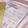 A close-up, angled shot of a transparent acrylic wedding invitation resting on a textured, light-colored fabric surface. The invitation features blue text and is partially overlaid by a yellow geometric design in the upper-left corner.