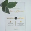 A transparent acrylic wedding invitation is displayed at an angle, leaning against a bouquet of soft green leaves resting over the top left corner.The invitation features elegant gold text.