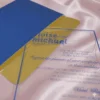 A transparent acrylic wedding invitation is shown partially tucked into a textured royal blue envelope. The invitation rests on a draped, light pink satin fabric that creates soft, shimmering reflections on the clear surface.