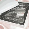 An angled, close-up view of an open white wedding invitation box resting on a soft, blush-toned fabric. The box features: A thick, clear acrylic invitation plaque** with a decorative scalloped edge, nestled inside on a black satin lining. Bold white typography on the acrylic card that reads "JOIN US FOR TWO FUN-FILLED DAYS ON THE BEACH" for a celebration on Location details for the "Round Rock Hotel Pompton" and RSVP information are clearly displayed at the bottom. The white lid of the box is partially visible, leaning against the base to reveal the luxury contents inside. The high-contrast design between the clear acrylic, white text, and dark lining creates a modern and high-end aesthetic.