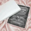 An angled, close-up view of an open white wedding invitation box resting on a soft, blush-toned fabric. The box features: A thick, clear acrylic invitation plaque** with a decorative scalloped edge, nestled inside on a black satin lining. Bold white typography on the acrylic card that reads "JOIN US FOR TWO FUN-FILLED DAYS ON THE BEACH" for a celebration on Location details for the "Round Rock Hotel Pompton" and RSVP information are clearly displayed at the bottom. The white lid of the box is partially visible, leaning against the base to reveal the luxury contents inside. The high-contrast design between the clear acrylic, white text, and dark lining creates a modern and high-end aesthetic.