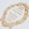 A oval-shaped transparent acrylic wedding invitation with a highly decorative, laser-cut edge. The card is framed by an intricate gold foil baroque border featuring ornate scrolls and flourishes.