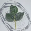 A clear acrylic wedding invitation in a geometric, diamond-like shape is positioned over a green leaf on a white surface. The invitation features the names in a large, bold sans-serif font, with the rest of the text in a smaller, lighter font. The text on the invitation read.
