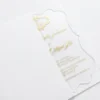 A transparent acrylic wedding invitation is shown partially tucked into a white paper pocket or sleeve. The image is shot at an angle on a white surface, highlighting the clarity of the invitation and the clean, minimalist presentation.
