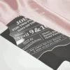 A transparent acrylic invitation is shown resting on a draped, shimmering pink satin fabric. The invitation is partially inserted into a white, pointed-flap envelope. The invitation features a unique, scalloped and wavy edge and contains white printed text. The design details include.