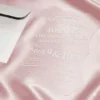 A transparent acrylic invitation is shown resting on a draped, shimmering pink satin fabric. The invitation is partially inserted into a white, pointed-flap envelope. The invitation features a unique, scalloped and wavy edge and contains white printed text. The design details include.