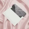 A transparent acrylic invitation is shown resting on a draped, shimmering pink satin fabric. The invitation is partially inserted into a white, pointed-flap envelope. The invitation features a unique, scalloped and wavy edge and contains white printed text. The design details include.