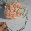 A person's hand holds a clear, geometric acrylic wedding invitation shaped like a multi-faceted diamond or gem. The invitation features an intricate gold border and gold lettering. In the background, out of focus, is a bouquet of light pink and peach roses.