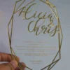 A hand holds a transparent acrylic wedding invitation with a striking laser-cut geometric gold border. The invitation is hexagonal in its overall silhouette, featuring sharp, intersecting gold lines that create a crystalline frame.