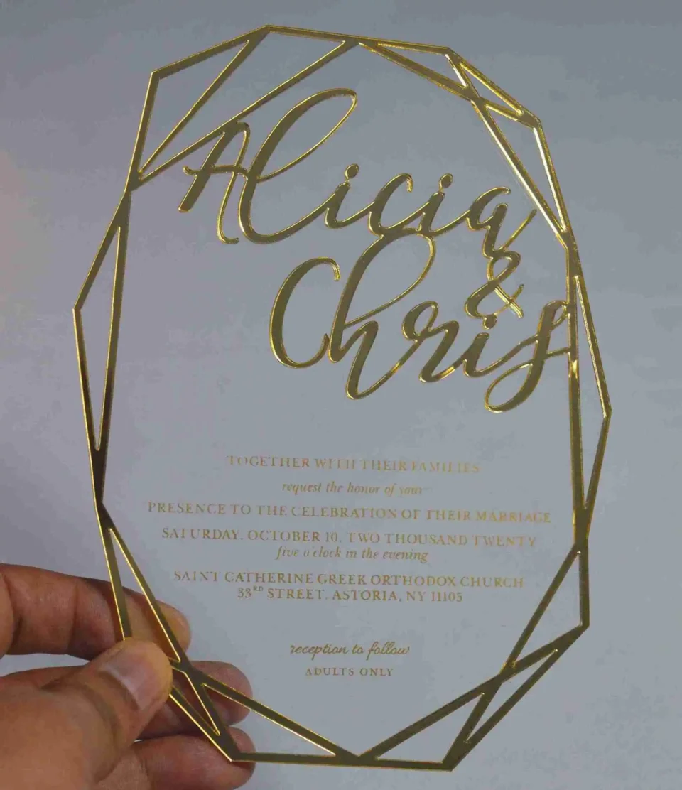 A hand holds a transparent acrylic wedding invitation with a striking laser-cut geometric gold border. The invitation is hexagonal in its overall silhouette, featuring sharp, intersecting gold lines that create a crystalline frame.