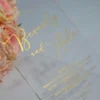 An transparent acrylic wedding invitation resting on a white surface. The card is leaning against a bouquet of soft peach and pink roses. The invitation features minimalist gold foil text with details.