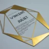 A clear acrylic wedding invitation in a sharp, geometric diamond shape lies flat on a light gray surface. The invitation features a striking border made of metallic gold triangles and intersecting lines that frame the central text.