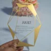 A transparent acrylic wedding invitation with a striking gold geometric frame is displayed leaning against a bouquet of soft peach and pink roses. The frame features sharp, crystalline lines and solid gold triangular accents in the corners.
