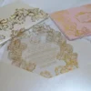An ornate wedding invitation suite is displayed on a white surface, featuring ivory, pink, and transparent acrylic elements decorated with elaborate gold foil filigree.