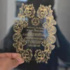 A hand holds up a transparent acrylic wedding invitation with an intricate, laser-cut scalloped edge. The invitation is decorated with a heavy gold foil filigree border of swirling acanthus leaves that frame the central text.