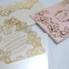 An ornate wedding invitation suite is displayed on a white surface, featuring ivory, pink, and transparent acrylic elements decorated with elaborate gold foil filigree.