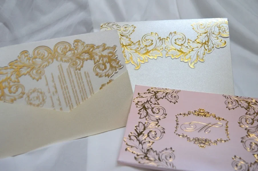 An ornate wedding invitation suite is displayed on a white surface, featuring ivory, pink, and transparent acrylic elements decorated with elaborate gold foil filigree.