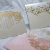 An ornate wedding invitation suite is displayed on a white surface, featuring ivory, pink, and transparent acrylic elements decorated with elaborate gold foil filigree.