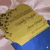 This image features a transparent acrylic wedding invitation with a uniquely shaped, scalloped silhouette. The card is displayed at an angle, resting on shimmering, light-pink satin fabric and partially tucked into a royal blue envelope.