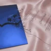 This image features a transparent acrylic wedding invitation with a uniquely shaped, scalloped silhouette. The card is displayed at an angle, resting on shimmering, light-pink satin fabric and partially tucked into a royal blue envelope.