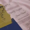 This image features a transparent acrylic wedding invitation with a uniquely shaped, scalloped silhouette. The card is displayed at an angle, resting on shimmering, light-pink satin fabric and partially tucked into a royal blue envelope.