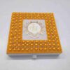 A high-angle, centered shot of a square **white wedding invitation box** featuring a vibrant and ornate lid design. The box is characterized by: * A thick, rectangular border encrusted with **bright gold-colored pearls** and small, shimmering **clear crystals** arranged in a repetitive, floral-like pattern. * A central square of smooth white fabric featuring a **custom gold monogram** with the letters "J" and "J" inside a circular, white and gold baroque-style crest. * The outermost edges of the border are finished with rows of tiny, uniform gold beads. * A small white satin pull-tab is visible on the front edge of the box. * The entire piece is presented against a neutral, light grey background, making the bold gold accents and intricate textures the primary focus.