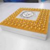 A high-angle, centered shot of a square **white wedding invitation box** featuring a vibrant and ornate lid design. The box is characterized by: * A thick, rectangular border encrusted with **bright gold-colored pearls** and small, shimmering **clear crystals** arranged in a repetitive, floral-like pattern. * A central square of smooth white fabric featuring a **custom gold monogram** with the letters "J" and "J" inside a circular, white and gold baroque-style crest. * The outermost edges of the border are finished with rows of tiny, uniform gold beads. * A small white satin pull-tab is visible on the front edge of the box. * The entire piece is presented against a neutral, light grey background, making the bold gold accents and intricate textures the primary focus.