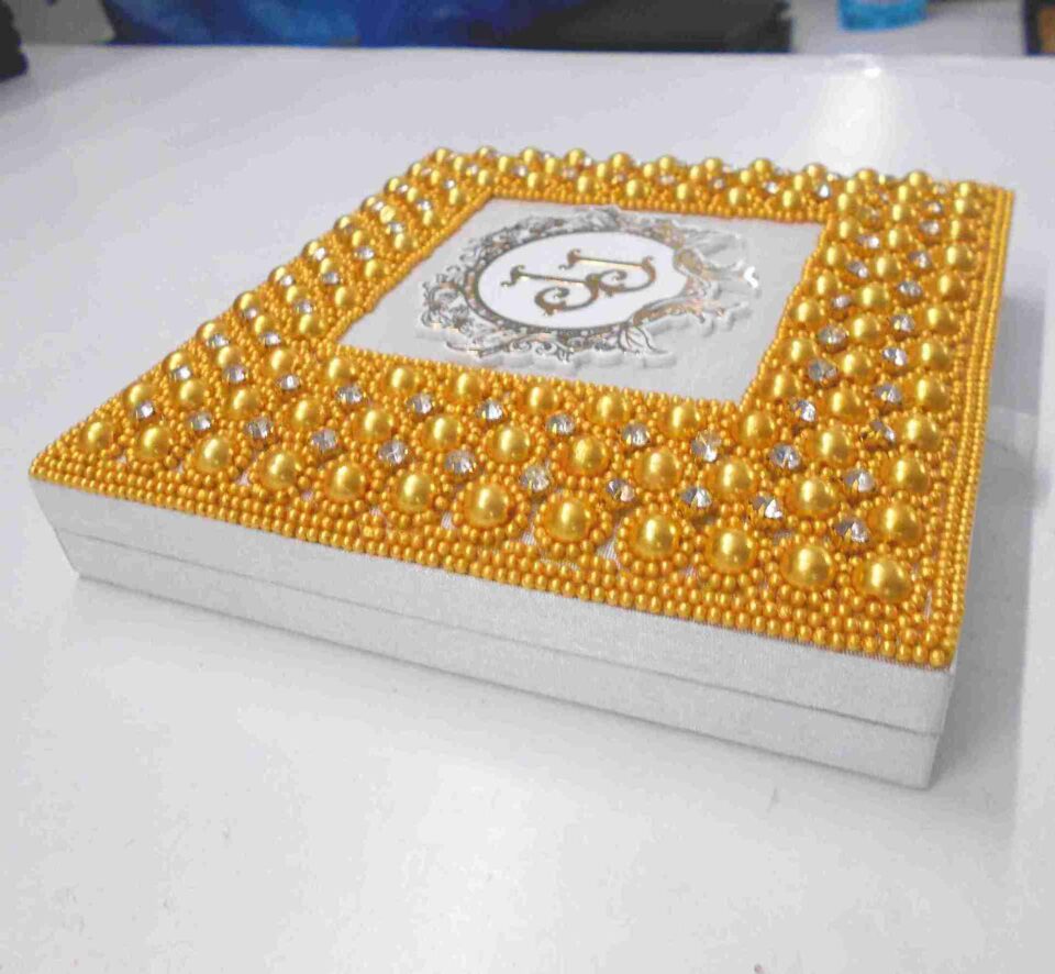 A high-angle, centered shot of a square **white wedding invitation box** featuring a vibrant and ornate lid design. The box is characterized by: * A thick, rectangular border encrusted with **bright gold-colored pearls** and small, shimmering **clear crystals** arranged in a repetitive, floral-like pattern. * A central square of smooth white fabric featuring a **custom gold monogram** with the letters "J" and "J" inside a circular, white and gold baroque-style crest. * The outermost edges of the border are finished with rows of tiny, uniform gold beads. * A small white satin pull-tab is visible on the front edge of the box. * The entire piece is presented against a neutral, light grey background, making the bold gold accents and intricate textures the primary focus.