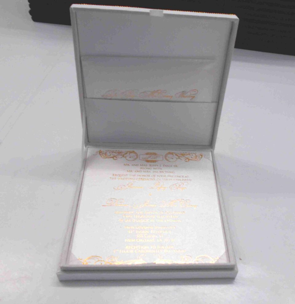 An open, **luxurious white wedding invitation box** featuring a formal stationery suite with gold accents. The interior includes: * A **white invitation card** in the base, printed with elegant **gold foil calligraphy** and framed by intricate gold scrollwork flourishes at the top and bottom. * The **inner lid** features a horizontal pocket holding a matching white card, also featuring a gold script heading. * The box is lined with a smooth, white fabric that gives it a clean and high-end appearance. * The top edge of the lid shows a hint of the **gold pearl-encrusted exterior**. * Set against a neutral, light grey background, the focus remains on the bright white textures and reflective gold detailing of the suite.