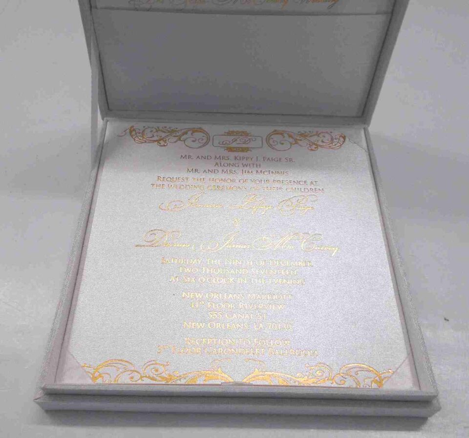 An open, **luxurious white wedding invitation box** featuring a formal stationery suite with gold accents. The interior includes: * A **white invitation card** in the base, printed with elegant **gold foil calligraphy** and framed by intricate gold scrollwork flourishes at the top and bottom. * The **inner lid** features a horizontal pocket holding a matching white card, also featuring a gold script heading. * The box is lined with a smooth, white fabric that gives it a clean and high-end appearance. * The top edge of the lid shows a hint of the **gold pearl-encrusted exterior**. * Set against a neutral, light grey background, the focus remains on the bright white textures and reflective gold detailing of the suite.