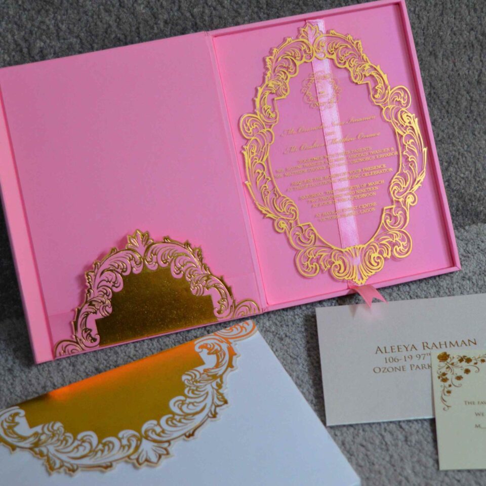 An angled close-up view of a luxury clear acrylic wedding invitation and coordinating stationery, set against a textured surface. The highlight of the suite is: An oval-shaped clear acrylic invitation featuring an intricate, laser-etched gold scrollwork border. The formal event details are printed in gold at the center of the plaque. The invitation rests partially atop a matching white invitation box with a gold-bordered lid. In the foreground, a gold-foiled RSVP card** is visible, featuring the initials and elegant floral accents in shimmering gold. In the background, a pink invitation folio with a gold central monogram adds a soft pop of color to the arrangement. The scene is captured in natural sunlight, creating soft shadows and emphasizing the high-shine, metallic finish of the gold elements.