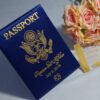 A high-angle view of a travel-themed wedding invitation set resting on a grey surface, featuring a royal blue silk passport invitation and a clear acrylic boarding pass. The suite includes:The Passport:Crafted from shimmering blue silk, it features the word "PASSPORT" and a large, intricate gold-foiled crest at the center. The names "**Rannice and Alphaus" and the date are printed at the bottom in elegant gold script.The Boarding Pass: A clear acrylic card designed to look like a flight ticket for "**All Eyez On Arrington Airways**." It features gold-etched details, including a barcode and the destination Decorative Accents: A cluster of soft peach-colored roses is placed in the background, adding a romantic contrast to the bold blue and gold stationery. The combination of luxury materials and a creative travel theme results in a unique and sophisticated invitation presentation.