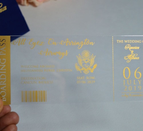 Inside Lid: A recessed panel holding a small white card with a gold-foiled monogram of the letters inside a floral wreath. The Base: A clear acrylic invitation card featuring an intricate, etched gold scrollwork border. The event details are printed in gold at the center. Foreground: A coordinating white RSVP card with shimmering gold foil text and decorative accents. Background: A gold-mirrored acrylic invitation is partially visible, reflecting the surrounding light and adding a high-shine element to the set.