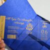 A high-angle view of a travel-themed wedding invitation set resting on a grey surface, featuring a royal blue silk passport invitation and a clear acrylic boarding pass. The suite includes:The Passport:Crafted from shimmering blue silk, it features the word "PASSPORT" and a large, intricate gold-foiled crest at the center. The names "**Rannice and Alphaus" and the date are printed at the bottom in elegant gold script.The Boarding Pass: A clear acrylic card designed to look like a flight ticket for "**All Eyez On Arrington Airways**." It features gold-etched details, including a barcode and the destination Decorative Accents: A cluster of soft peach-colored roses is placed in the background, adding a romantic contrast to the bold blue and gold stationery. The combination of luxury materials and a creative travel theme results in a unique and sophisticated invitation presentation.
