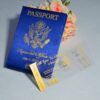 A high-angle view of a travel-themed wedding invitation set resting on a grey surface, featuring a royal blue silk passport invitation and a clear acrylic boarding pass. The suite includes:The Passport:Crafted from shimmering blue silk, it features the word "PASSPORT" and a large, intricate gold-foiled crest at the center. The names "**Rannice and Alphaus" and the date are printed at the bottom in elegant gold script.The Boarding Pass: A clear acrylic card designed to look like a flight ticket for "**All Eyez On Arrington Airways**." It features gold-etched details, including a barcode and the destination Decorative Accents: A cluster of soft peach-colored roses is placed in the background, adding a romantic contrast to the bold blue and gold stationery. The combination of luxury materials and a creative travel theme results in a unique and sophisticated invitation presentation.