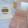 A high-angle shot featuring an open peach-colored wedding invitation box set on a white surface with draped white fabric and a gold lace cuff. On the left, the closed lid of the box displays a central gold plaque with the monogram "R & P," framed by purple scrollwork accented with pearls. On the right, the open box base reveals a formal acrylic invitation for "James Parker & Karen Brett" printed with gold text. A gold RSVP card with matching scrollwork is partially tucked into a pocket in the open lid.