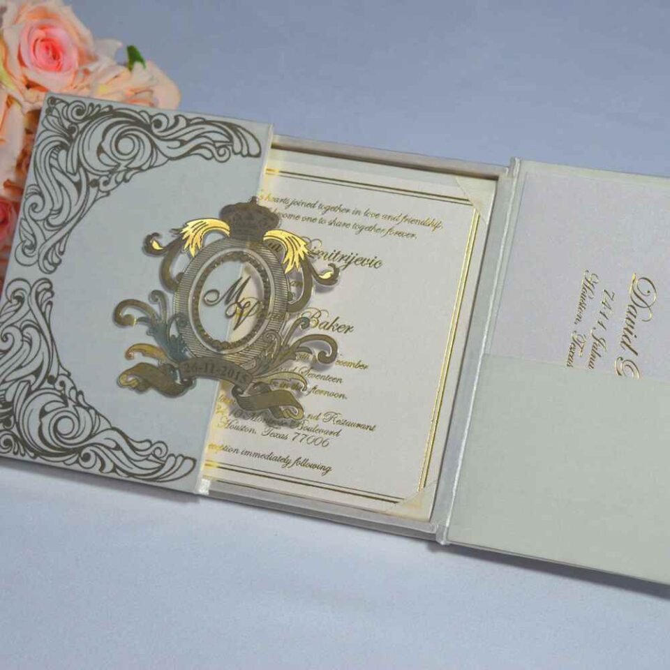 An open, ivory silk gatefold invitation box revealing a formal wedding invitation for Mia Dimitrijevic and David Baker. The invitation features elegant gold script and a gold-stamped circular monogram on the left side. The card is framed with a thin gold border. The left flap of the box contains a pocket holding a matching RSVP card. A bouquet of soft peach-colored roses sits in the background on a light grey surface.