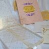 A high-angle shot featuring an open peach-colored wedding invitation box set on a white surface with draped white fabric and a gold lace cuff. On the left, the closed lid of the box displays a central gold plaque with the monogram "R & P," framed by purple scrollwork accented with pearls. On the right, the open box base reveals a formal acrylic invitation for "James Parker & Karen Brett" printed with gold text. A gold RSVP card with matching scrollwork is partially tucked into a pocket in the open lid.