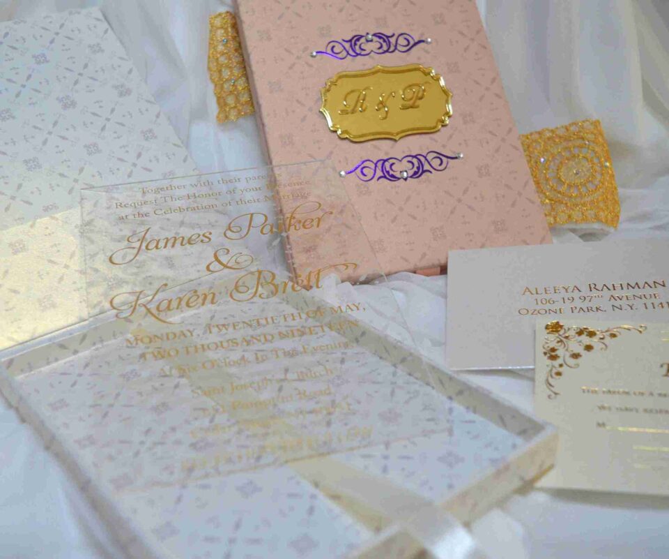 A high-angle shot featuring an open peach-colored wedding invitation box set on a white surface with draped white fabric and a gold lace cuff. On the left, the closed lid of the box displays a central gold plaque with the monogram "R & P," framed by purple scrollwork accented with pearls. On the right, the open box base reveals a formal acrylic invitation for "James Parker & Karen Brett" printed with gold text. A gold RSVP card with matching scrollwork is partially tucked into a pocket in the open lid.