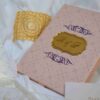 A rectangular light pink invitation box with a subtle geometric pattern resting on draped white fabric. The lid features a central gold-mirrored plaque engraved with the monogram "R & L," flanked by purple scrollwork designs accented with small pearls. A decorative gold lace cuff and a partially visible white card with the name "Aleeya" are placed nearby.