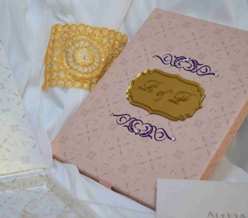 A rectangular light pink invitation box with a subtle geometric pattern resting on draped white fabric. The lid features a central gold-mirrored plaque engraved with the monogram "R & L," flanked by purple scrollwork designs accented with small pearls. A decorative gold lace cuff and a partially visible white card with the name "Aleeya" are placed nearby.
