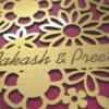 A high-angle shot of a square, chocolate-brown invitation box. The lid features an intricate, laser-cut gold floral pattern of various daisies and blossoms over a vibrant magenta background. A gold banner integrated into the floral design is engraved with the names "Aakash & Preeti" in an elegant script. A second, identical box is partially visible in the blurred background.