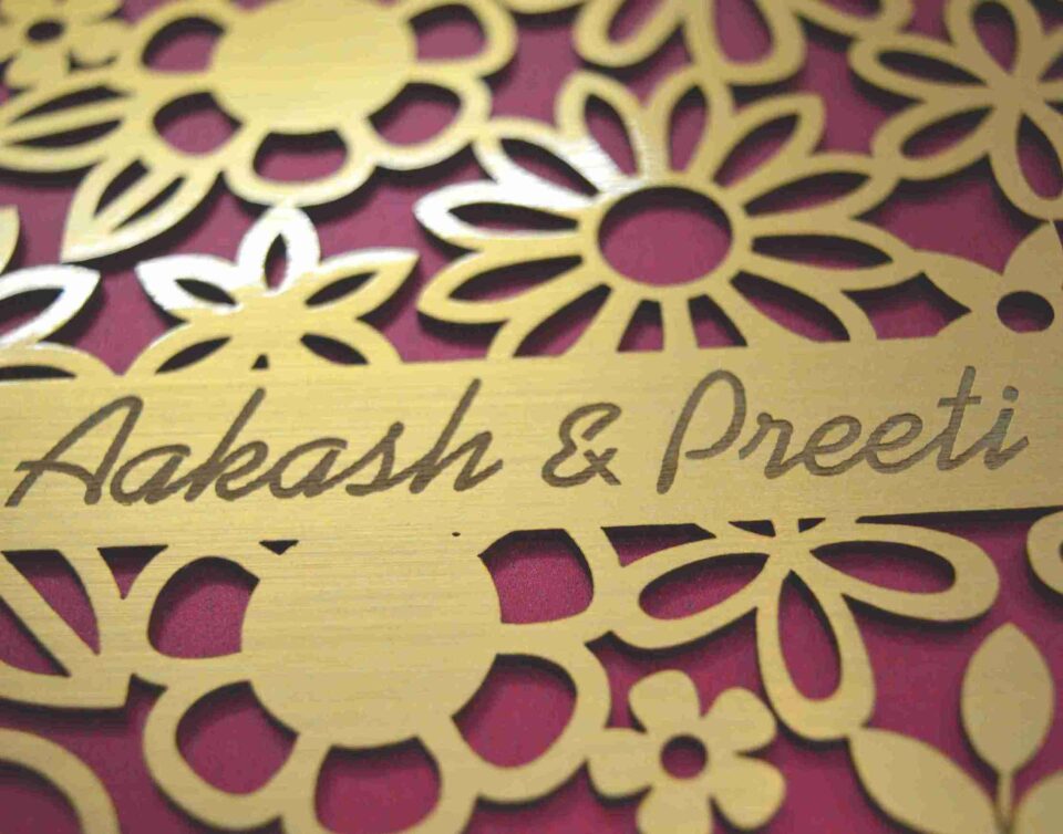 A high-angle shot of a square, chocolate-brown invitation box. The lid features an intricate, laser-cut gold floral pattern of various daisies and blossoms over a vibrant magenta background. A gold banner integrated into the floral design is engraved with the names "Aakash & Preeti" in an elegant script. A second, identical box is partially visible in the blurred background.