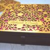 A high-angle shot of a square, chocolate-brown invitation box. The lid features an intricate, laser-cut gold floral pattern of various daisies and blossoms over a vibrant magenta background. A gold banner integrated into the floral design is engraved with the names "Aakash & Preeti" in an elegant script. A second, identical box is partially visible in the blurred background.